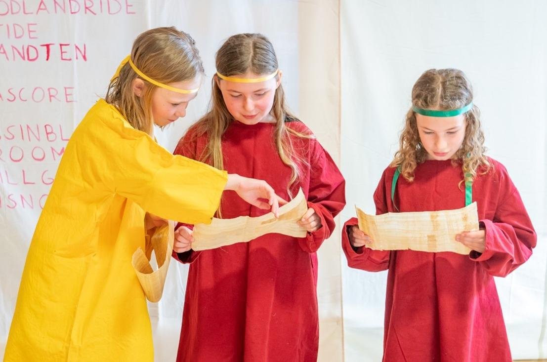 A photograph showing three girls dressed in Roman costume reading from two ancient scrolls and preparing a recitation